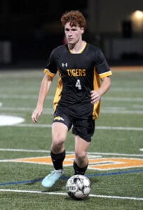 Soccer player in black Tigers jersey dribbles ball on field during night game.