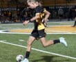Soccer player in black and yellow uniform dribbling ball on field at night game.