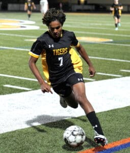Soccer player in black Tigers jersey dribbling a ball near sideline during a night game on a green field.