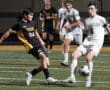 Two high school soccer players from opposing teams compete for the ball during a nighttime match on a grassy field.