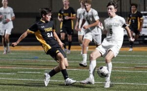 Two high school soccer players from opposing teams compete for the ball during a nighttime match on a grassy field.