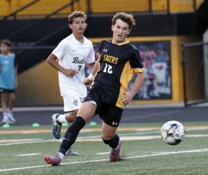 Soccer player in black and yellow uniform dribbling the ball on a field, pursued by an opponent.