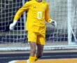Soccer goalie in yellow uniform prepares to kick the ball during a match on a turf field.