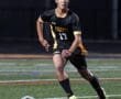 Soccer player in black and yellow uniform dribbles ball on field during night game.