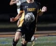 Soccer player in black Tigers uniform dribbles ball during night match on field, pursued by opponent.