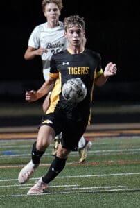 Soccer player in black Tigers uniform dribbles ball during night match on field, pursued by opponent.