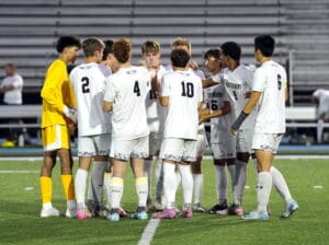 Youth soccer team huddles on field, wearing white uniforms, showing team spirit and camaraderie under stadium lights.