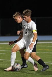Soccer match action, player in white jersey dribbling the ball against opponent in black at night.