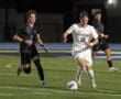 Soccer player in white jersey advancing the ball past opponent in black on a field during a night match.