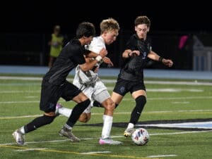 Soccer players in intense match, two in black attacking player in white on green field.