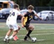 Boys' high school soccer match, player in black and yellow dribbling past opponent in white on the field.