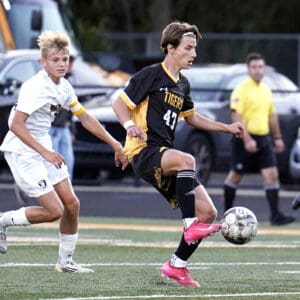 Teen soccer players in action during a match, one in a black uniform controlling the ball.