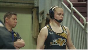 Female wrestler in uniform and headgear before a match, focused and standing beside coach near the stairs.