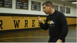 Man instructing in a wrestling gym, emphasizing technique, with motivational banners on the wall in the background.