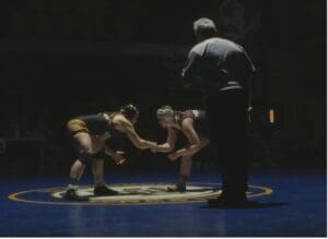 Two female wrestlers face off on a blue mat under spotlight, with a referee nearby, during a wrestling match.