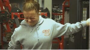 Person in gray hoodie exerting effort at the gym with weights, eyes closed in concentration.