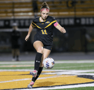 Female soccer player in black and yellow uniform dribbling a ball on a field at night.