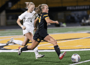 Two female soccer players compete for the ball during an intense match on a green and yellow field.