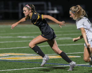 Two female soccer players sprinting during a competitive match on a well-lit field at night.