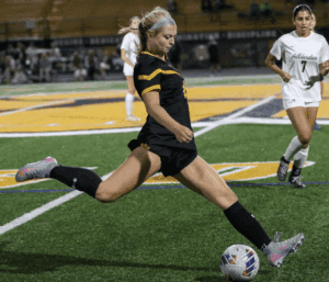 Soccer player in black kicks ball during match, while opponent in white observes on green field.