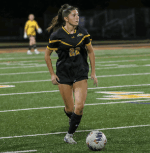 Soccer player in black jersey dribbling on field at night game, with a goalkeeper in the background.