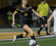 Female soccer player in black and gold uniform dribbles ball during night match on green field.