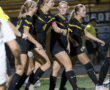 Soccer team celebrates a goal on the field, wearing black and yellow uniforms during a night match.