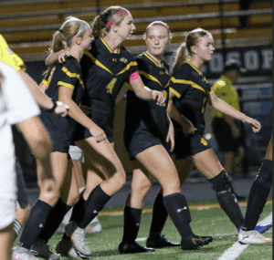 Soccer team celebrates a goal on the field, wearing black and yellow uniforms during a night match.