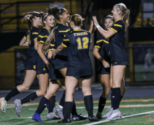 Girls' soccer team celebrating on the field in dark uniforms with yellow stripes.