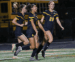 Young women's soccer team jubilant after scoring on field at night, wearing matching navy uniforms, celebrating victory.