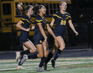 Young women's soccer team jubilant after scoring on field at night, wearing matching navy uniforms, celebrating victory.