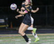 Female soccer player in black uniform controls the ball during a night match, wearing a captain's armband.