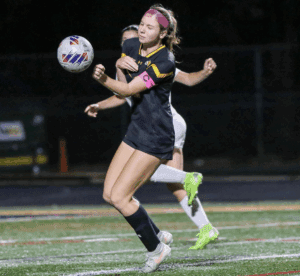 Female soccer player in black uniform controls the ball during a night match, wearing a captain's armband.