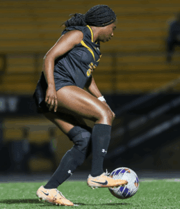 Soccer player in black jersey dribbling a ball on a field during a night game.