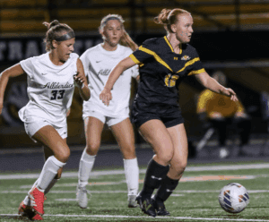 Girls' soccer match action, players in white and black jerseys compete for the ball on the field.