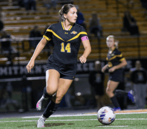 Soccer player in black jersey with pink captain armband dribbles ball on field during night match.