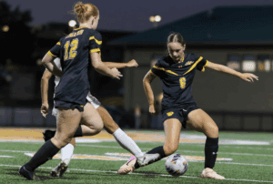 Two soccer players in black uniforms compete for the ball on a field during a night game.