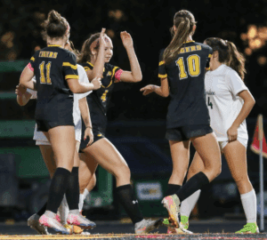 Soccer players celebrate a goal in a nighttime match, wearing black uniforms with yellow accents.