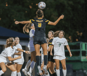 Soccer player in black jumps for header against team in white during intense match under evening lights.