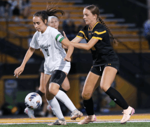 Two soccer players competing for the ball on the field during a match, one in white, one in black and yellow.