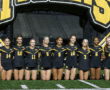 Girls' soccer team in black uniforms posing on field at night, under yellow-and-black inflatable arch.
