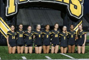 Girls' soccer team in black uniforms posing on field at night, under yellow-and-black inflatable arch.