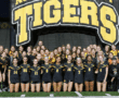 High school girls' soccer team poses under Tigers inflatable on the field, wearing black and yellow uniforms.