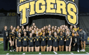 High school girls' soccer team poses under Tigers inflatable on the field, wearing black and yellow uniforms.