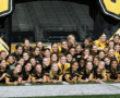 Girls soccer team in black and yellow uniforms poses cheerfully in front of inflatable arch on field.