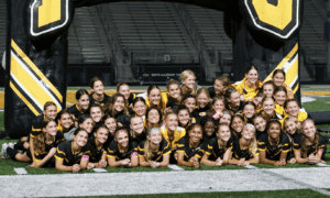 Girls soccer team in black and yellow uniforms poses cheerfully in front of inflatable arch on field.