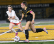 Two female soccer players in action on a field during a game, with one in a black uniform dribbling the ball.