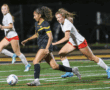 Women's soccer match, player in black dribbling ball, opponents in white pursuing on field at night.