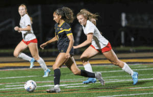 Women's soccer match, player in black dribbling ball, opponents in white pursuing on field at night.