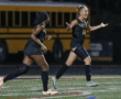 Two female soccer players celebrating on the field in front of a yellow school bus at night.